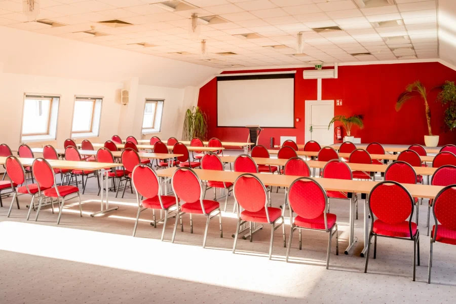 Saal mit roten St&uuml;hlen an hellen Tischen, an der roten Stirnwand eine gro&szlig;e Leinwand