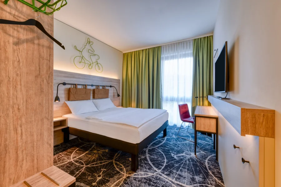 Hotelzimmer mit Doppelbett, gr&uuml;nen Vorh&auml;ngen und Fahrrad-Wandgrafik, rechts Schreibtisch mit rotem Stuhl und Fernseher