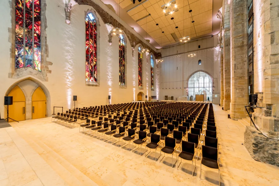 Kirchenschiff der Johanniskirche als Veranstaltungssaal: Reihenbestuhlung auf hellem Steinboden, bunte Kirchenfenster und Blick zur B&uuml;hne