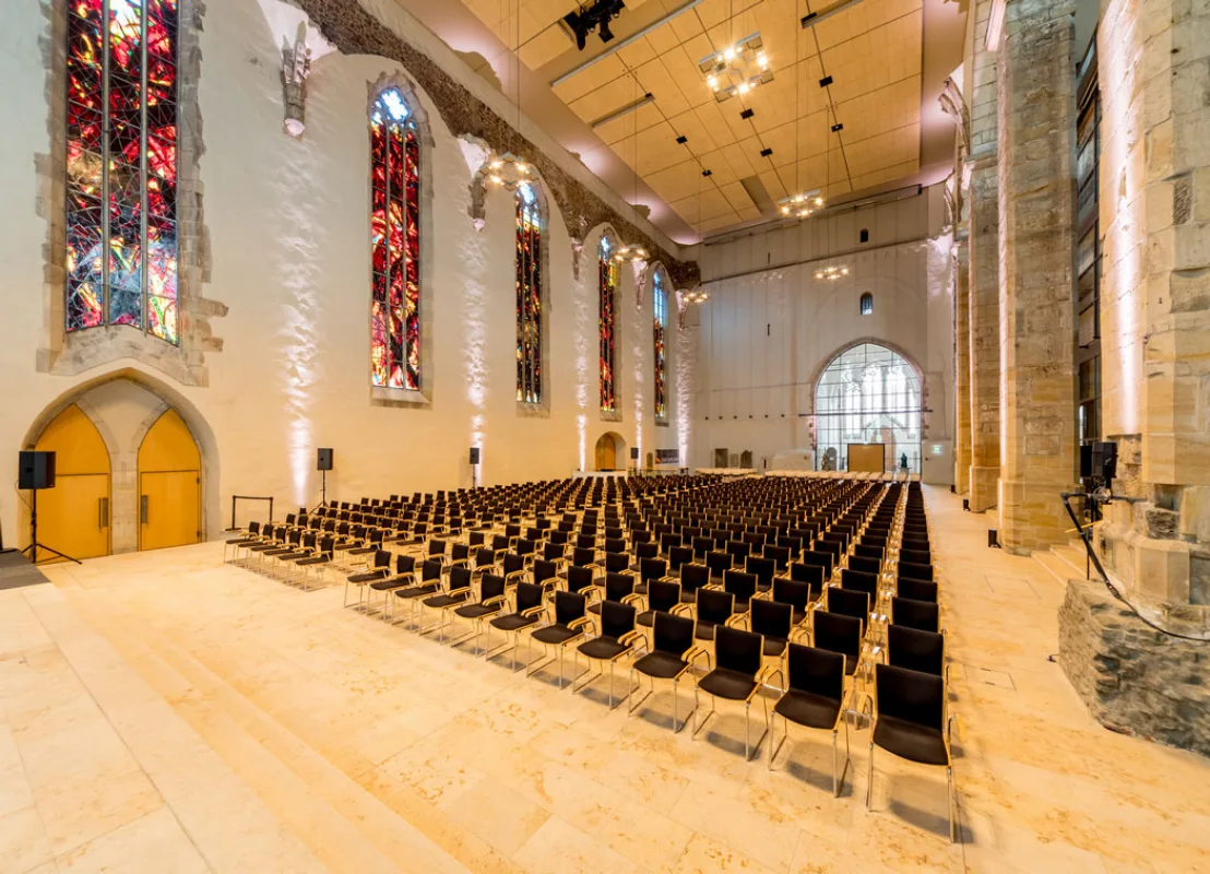 Kirchenschiff der Johanniskirche als Veranstaltungssaal: Reihenbestuhlung auf hellem Steinboden, bunte Kirchenfenster und Blick zur Bühne