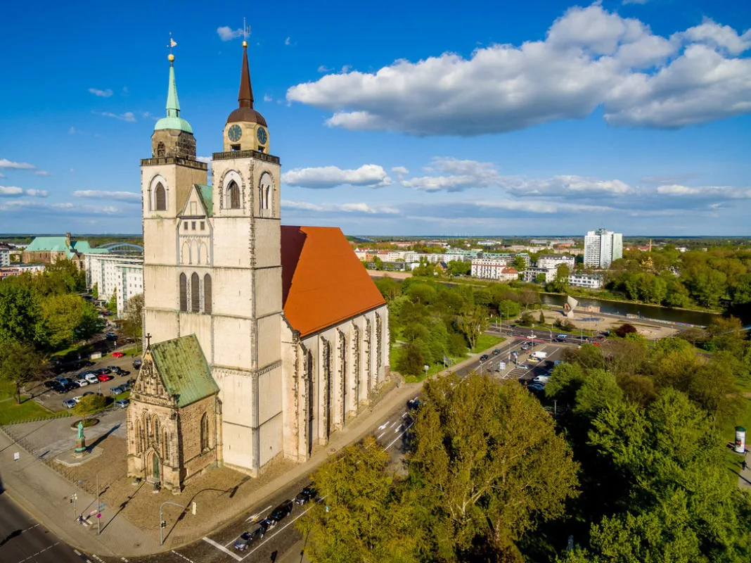 Luftbild der Johanniskirche mit rotem Dach und beiden Türmen, dahinter die Elbe und die Magdeburger Innenstadt
