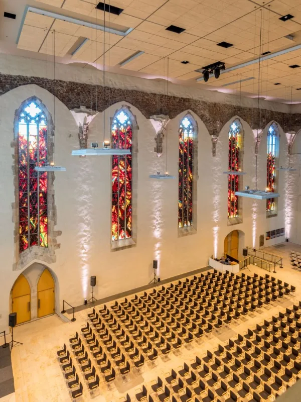 Saal der Johanniskirche von der Empore aus: hohe gotische Fenster mit rot leuchtendem Glas, weiß verputzte Wände und dicht gestellte Stuhlreihen