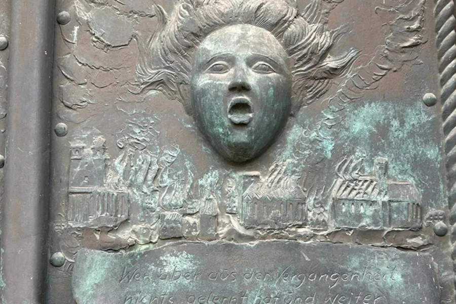 Bronzerelief an der Johanniskirchentuer zur Zerstoerung Magdeburgs 1631: schreiendes Gesicht mit wehendem Haar ueber der brennenden Stadtsilhouette