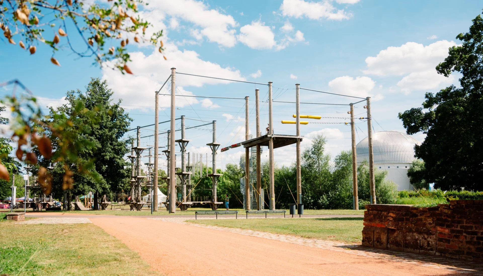 Kletterpark Magdeburg mit hoher Sprungplattform und Seilelementen unter blauem Wolkenhimmel, im Hintergrund die weisse Kuppel im Stadtpark Rotehorn