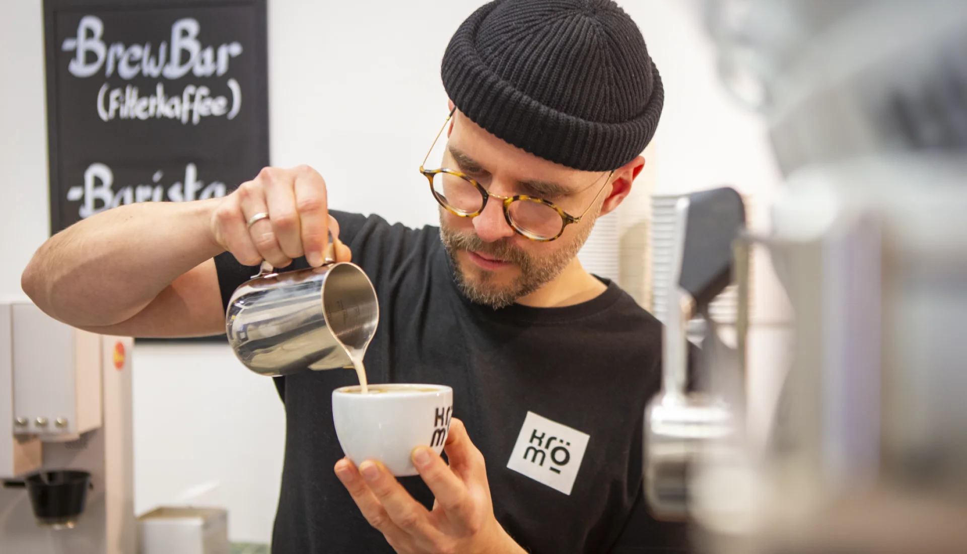 Barista der KR&Ouml;M Kaffeer&ouml;sterei mit dunkler M&uuml;tze und Brille gie&szlig;t Milchschaum in eine Tasse mit Logo, im Hintergrund Tafeln mit der Aufschrift Brew Bar