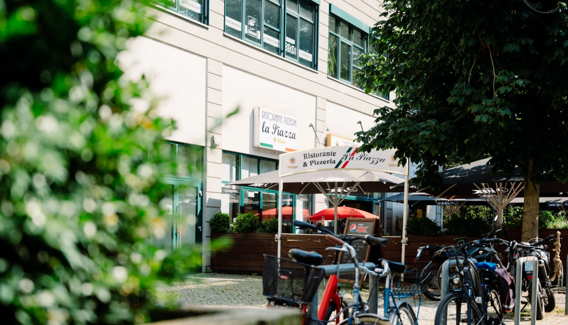 Terrasse des La Piazza unter gro&szlig;en dunklen Sonnenschirmen, mit Korbst&uuml;hlen und roten Holztischen zwischen Hecken und Lavendel