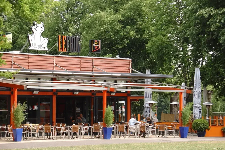 Terrasse des Restaurants Le Frog mit orangefarbenen Stuetzen, weissen Sonnenschirmen und besetzten Tischen, auf dem Dach der weisse Frosch-Schriftzug