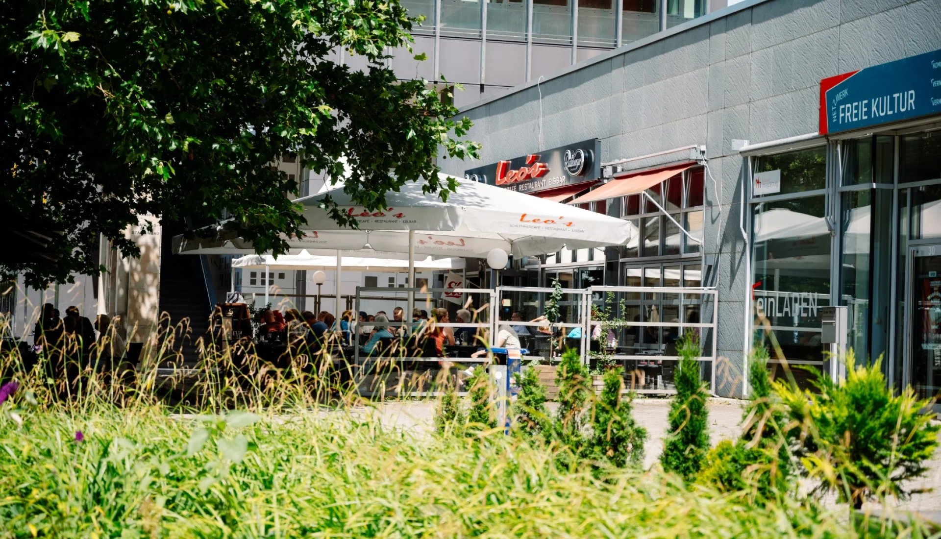 Gut besuchte Aussenterrasse von Leos Schlemmercafe unter weissen Sonnenschirmen mit rotem Leuchtschriftzug an der Fassade, vorn Graeser und junge Baeume
