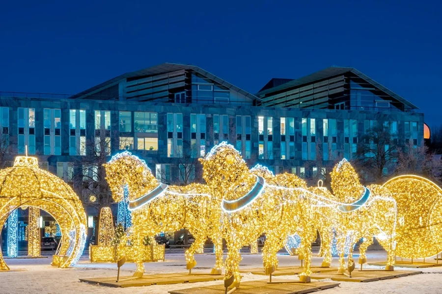 Lichterwelt Magdeburg: goldene Pferde aus Lichterketten ziehen einen leuchtenden Schlitten zwischen begehbaren Riesen-Weihnachtskugeln, dahinter ein modernes Buerogebaeude in der Daemmerung