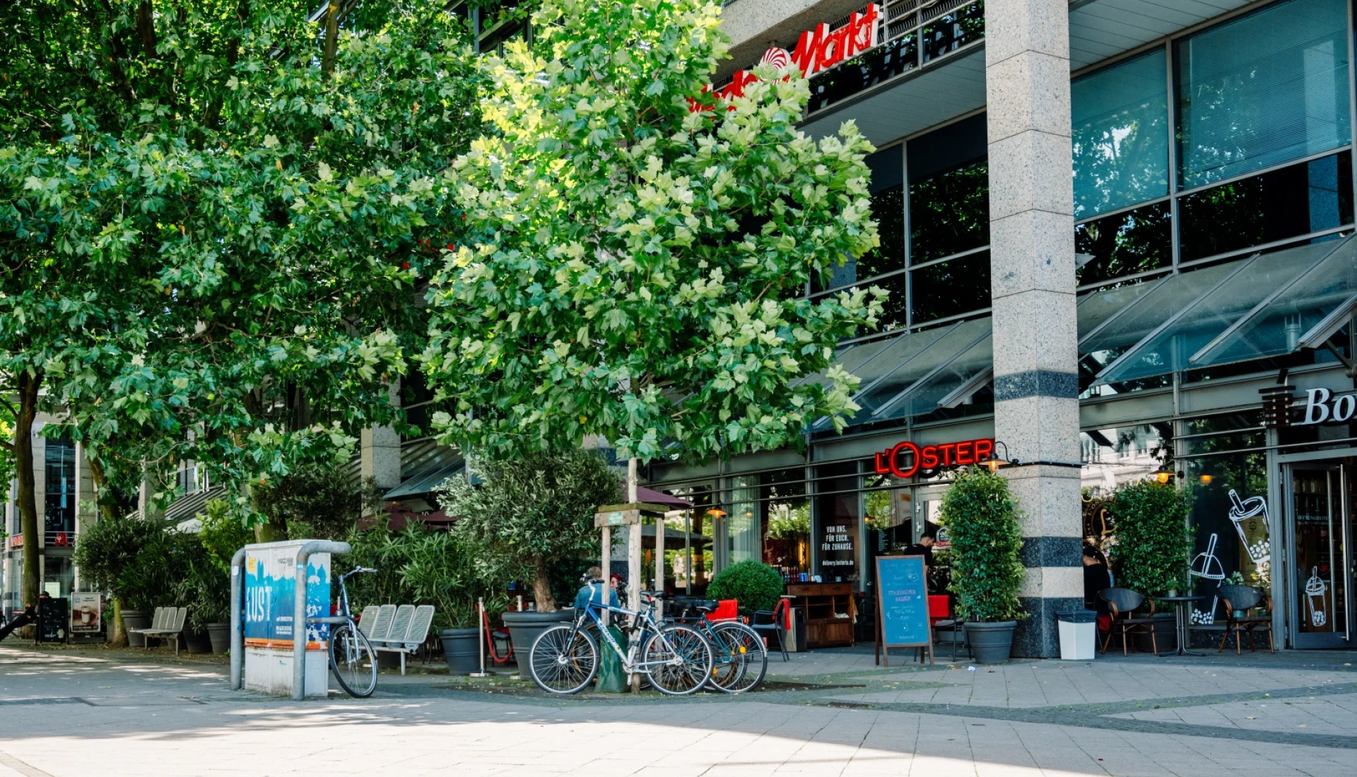Die L'Osteria im Erdgeschoss eines modernen Geschaeftshauses am Breiten Weg, Terrasse mit Baenken und Fahrradstaendern unter grossen Baeumen