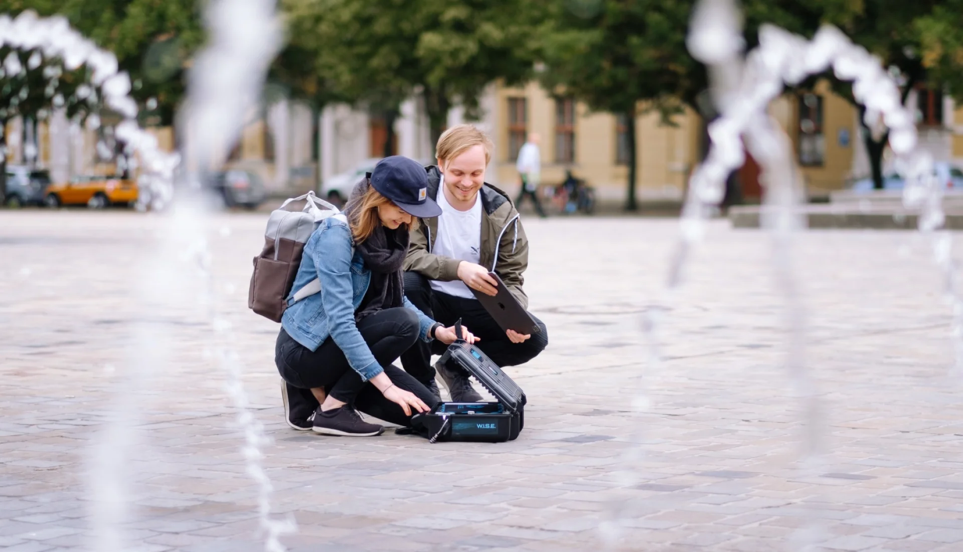 Zwei Teilnehmende einer Lukullus-Tour hocken zwischen Fontaenen auf dem Domplatz und oeffnen einen schwarzen Ausruestungskoffer mit der Aufschrift W.I.S.E.