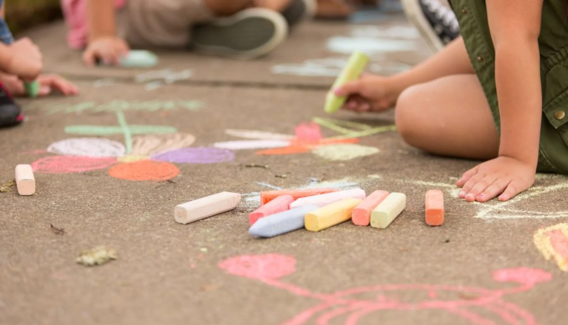 Kinder malen mit bunter Strassenkreide Blumen auf Asphalt, in der Mitte liegt ein Haufen Kreidestuecke in Rosa, Gelb, Blau und Orange