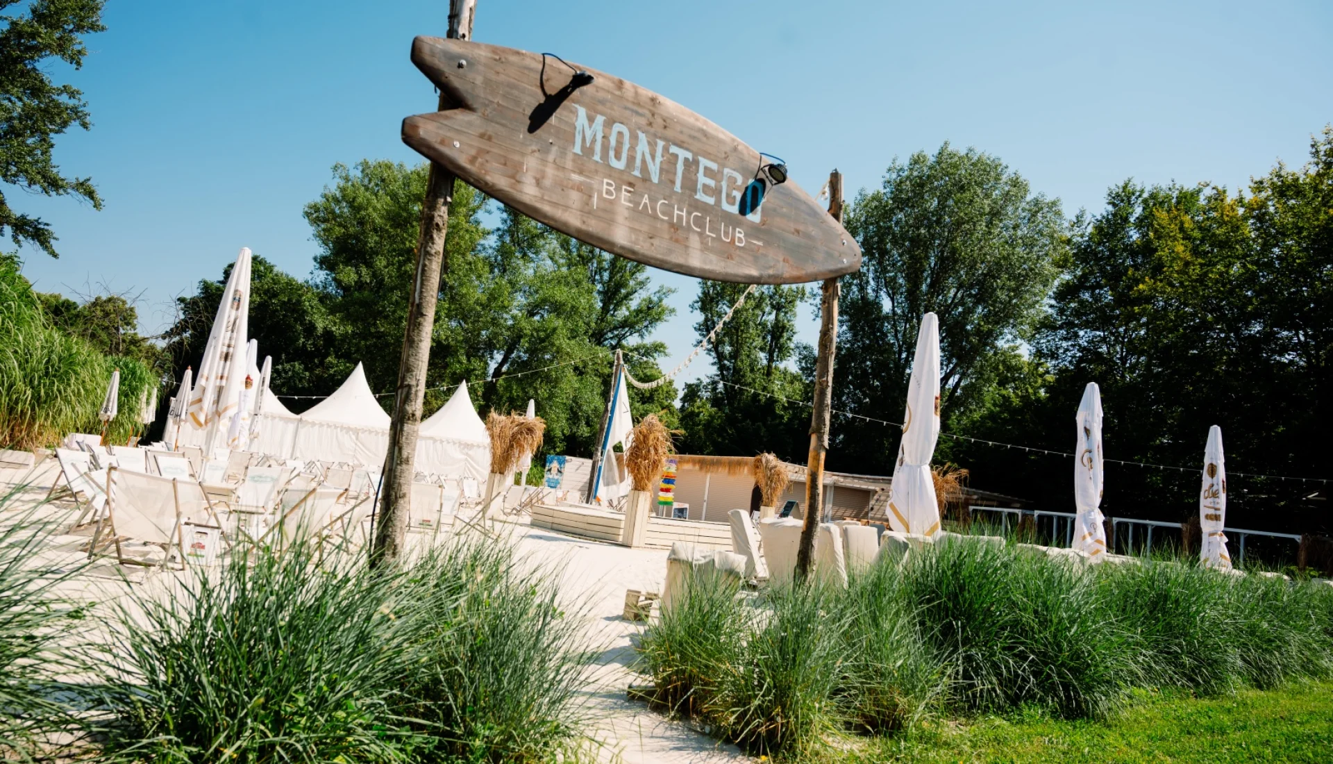 Surfbrett-Schild &bdquo;Montego Beachclub&ldquo; am Eingang zum Strand, dahinter helle Strandliegen, wei&szlig;e Zelte und aufgerollte Sonnenschirme im Sand