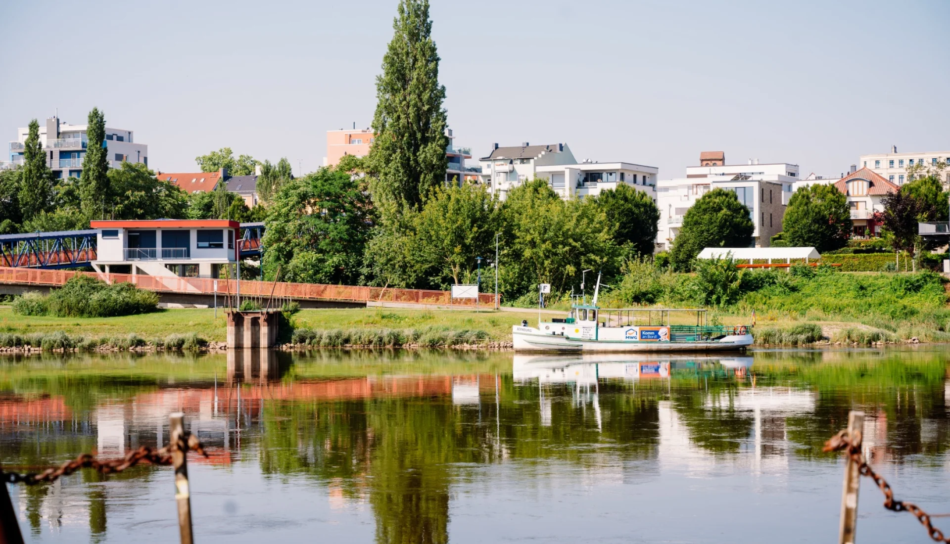 F&auml;hre Buckau an der Anlegestelle am gr&uuml;nen Elbufer vor modernen Wohnh&auml;usern, links eine rote Stahlbr&uuml;cke, das Ufer spiegelt sich im Wasser