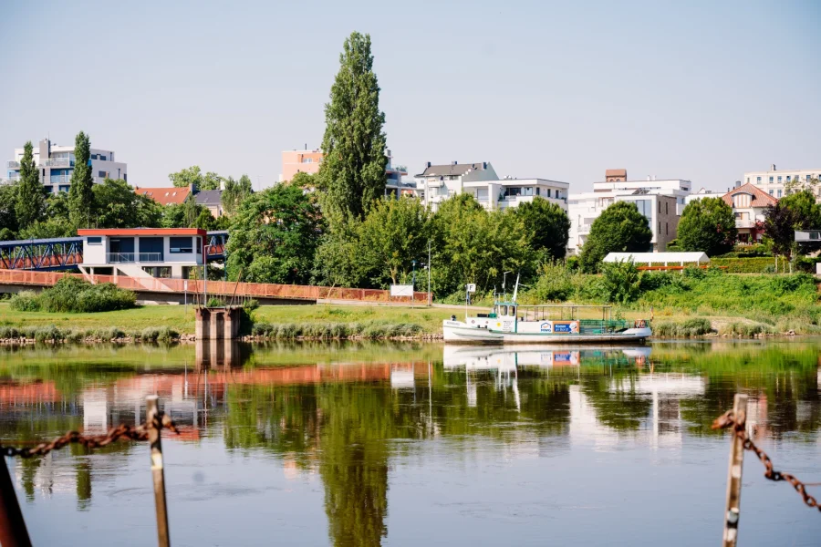 F&auml;hre Buckau an der Anlegestelle am gr&uuml;nen Elbufer vor modernen Wohnh&auml;usern, links eine rote Stahlbr&uuml;cke, das Ufer spiegelt sich im Wasser