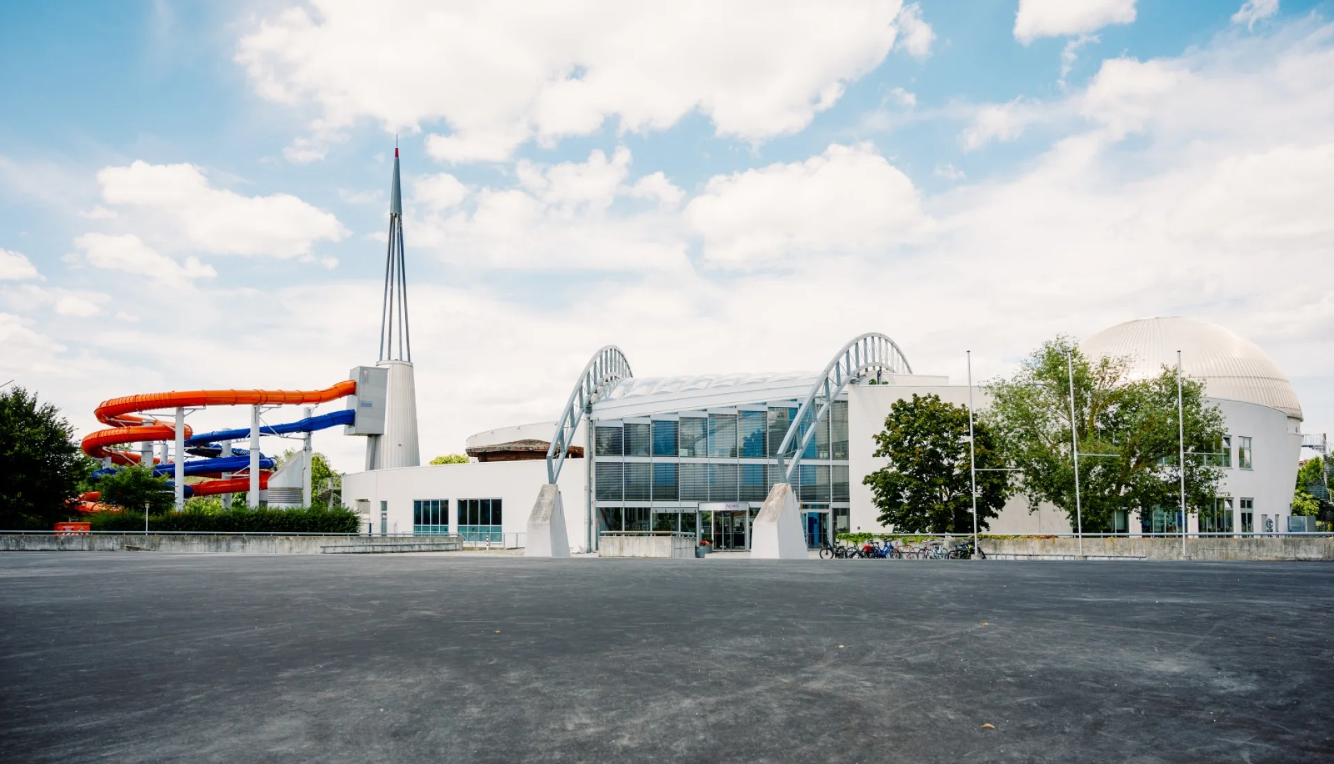 Au&szlig;enansicht des NEMO mit gl&auml;serner Eingangsfront unter zwei wei&szlig;en B&ouml;gen, rechts die Saunakuppel, links die bunten Rutschen
