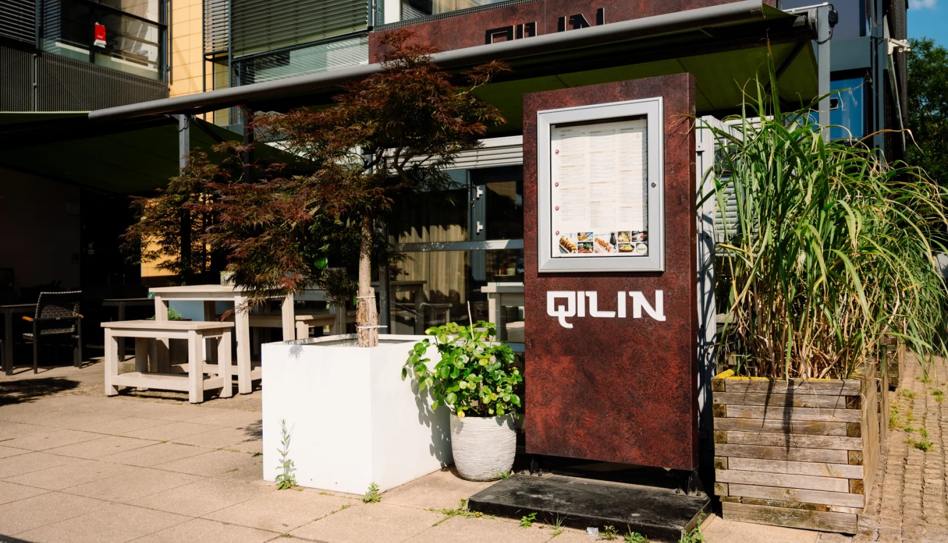 Eingangsbereich des QILIN mit rostroter Men&uuml;-Stele, hellen Holztischen auf der Terrasse, japanischem Ahorn und hohen Gr&auml;sern