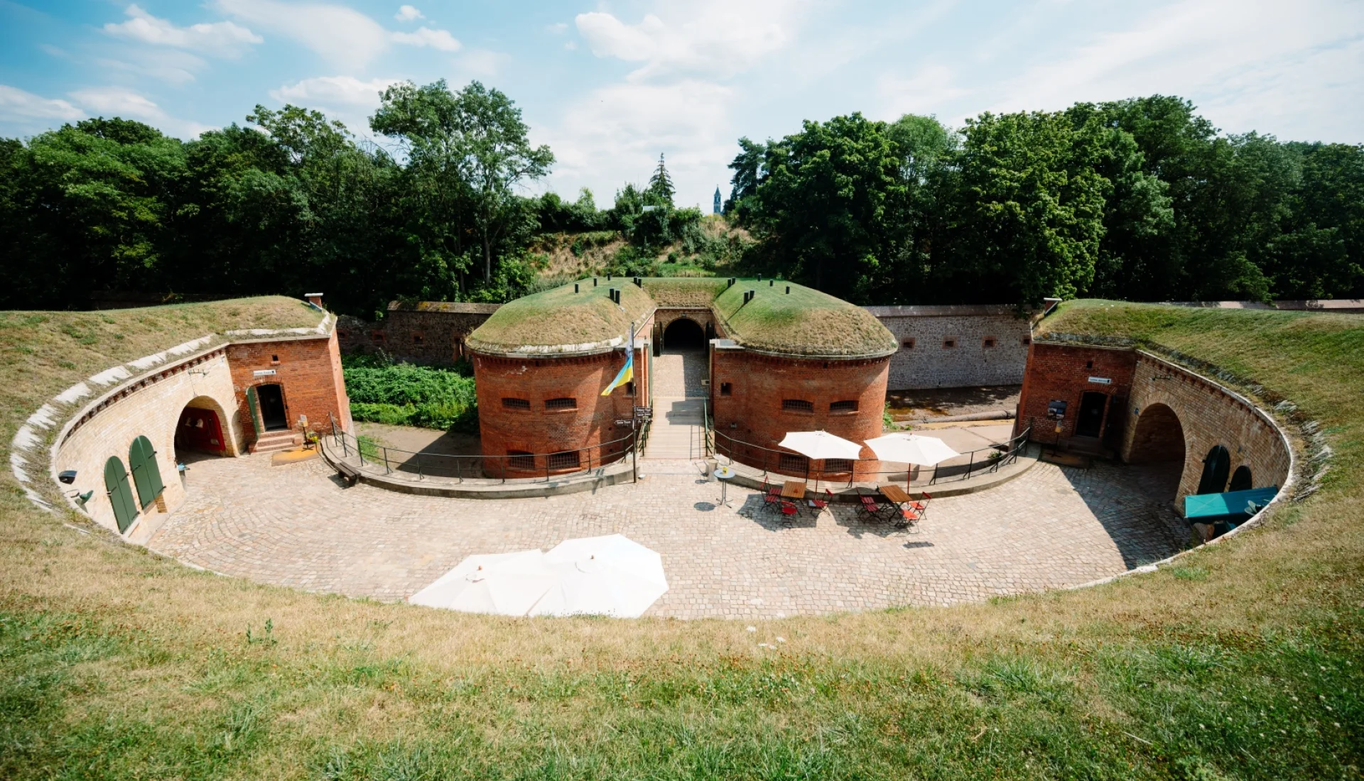 Ravelin 2 von oben: halbrunde Festungsanlage aus rotem Backstein mit grasbewachsenen D&auml;chern, im gepflasterten Hof wei&szlig;e Sonnenschirme und bunte St&uuml;hle
