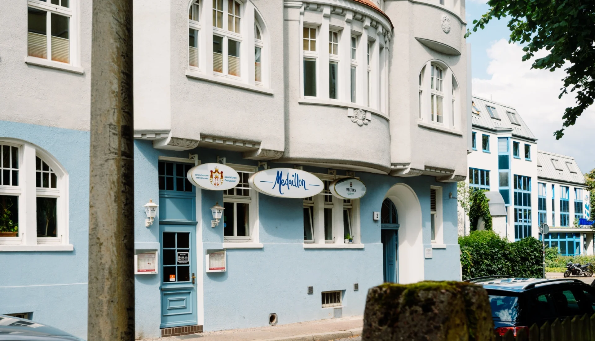 Hellblau-weisses Jugendstil-Eckhaus des Restaurants Medaillon: ovales Schild mit blauem Schriftzug ueber den Fenstern, blaue Tuer mit Laternen, davor Kopfsteinpflaster