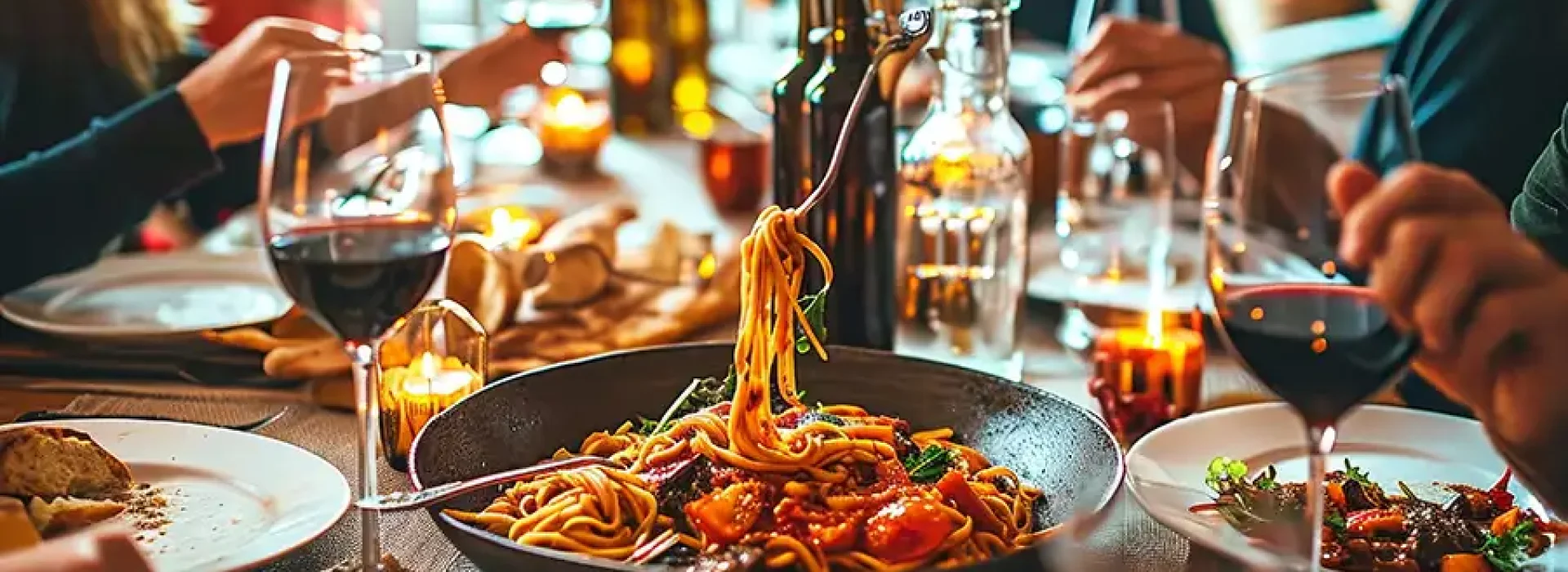 Gedeckter Tisch mit dampfender Pasta in schwarzer Pfanne, Rotweinglaesern und Brot, drumherum lachende Gaeste beim gemeinsamen Essen