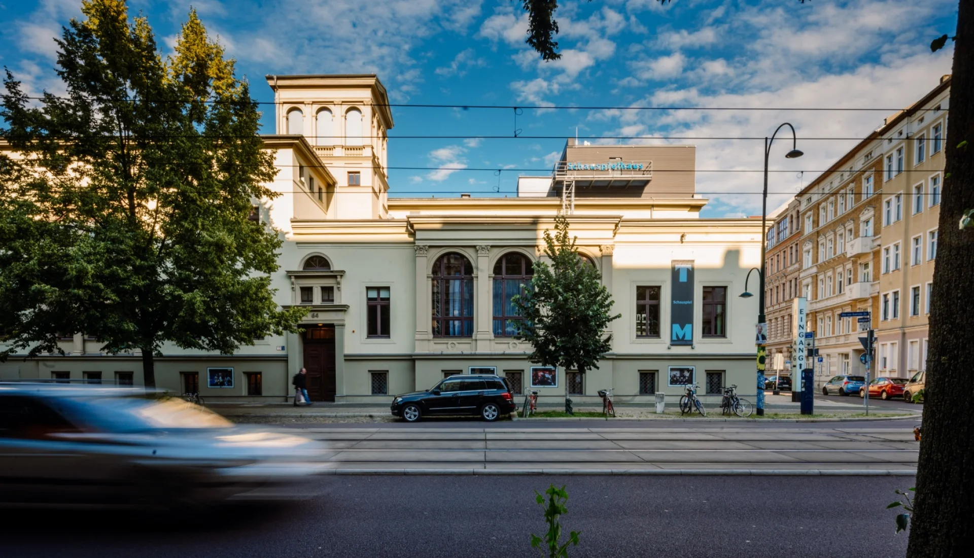 Schauspielhaus des Theaters Magdeburg von der Stra&szlig;e aus, mit blauem TM-Banner an der Fassade und einem vorbeifahrenden Auto