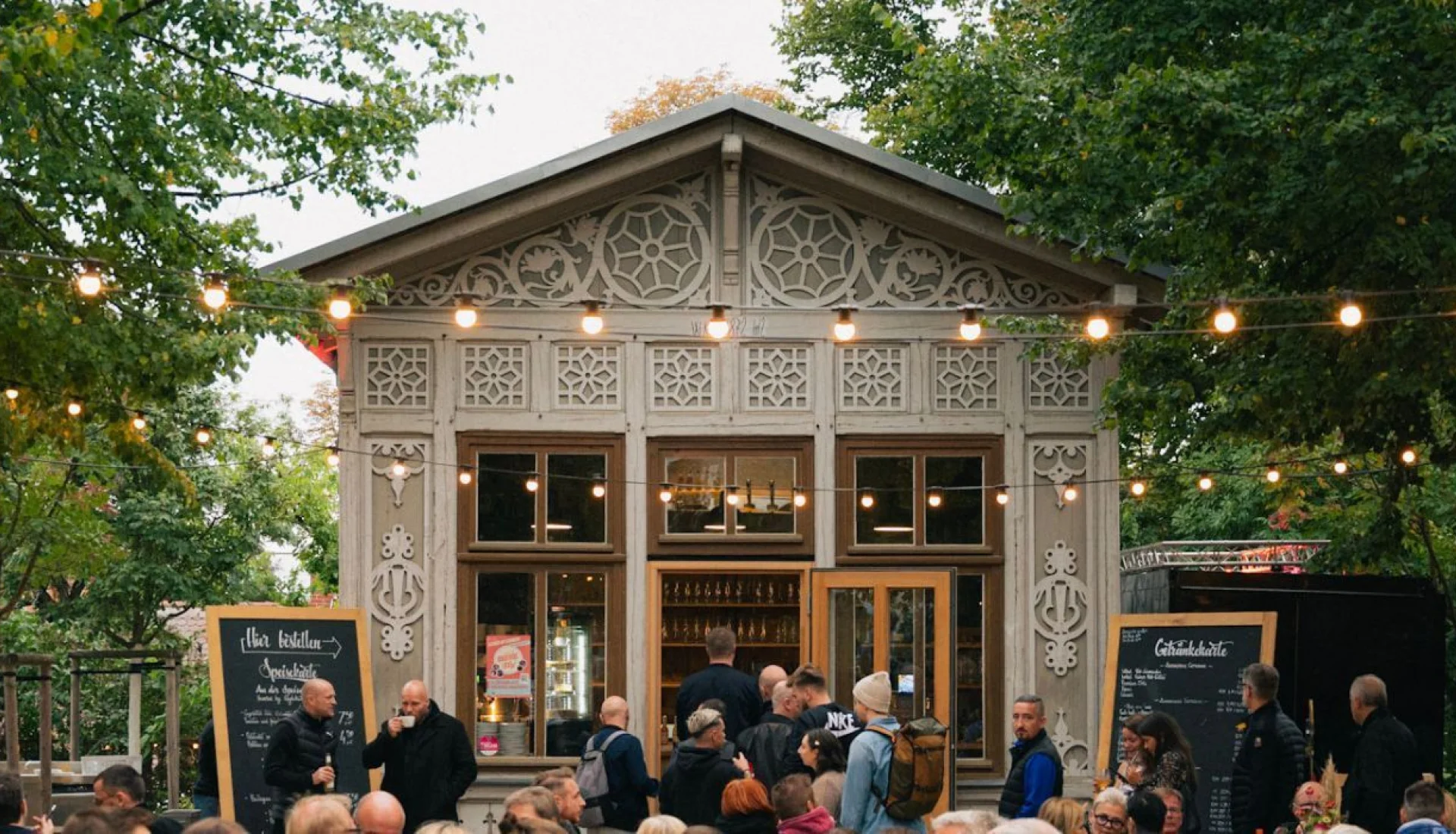 Wei&szlig;er Holzpavillon der Schweizer Milchkuranstalt mit geschnitzten Ornamenten und Lichterketten, davor G&auml;ste zwischen Kreidetafeln mit Speise- und Getr&auml;nkekarte
