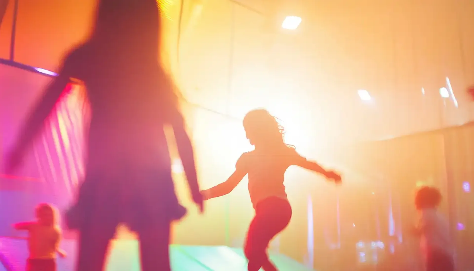 Kinder springen als Silhouetten durch einen bunt beleuchteten Trampolinpark in warmem Gegenlicht