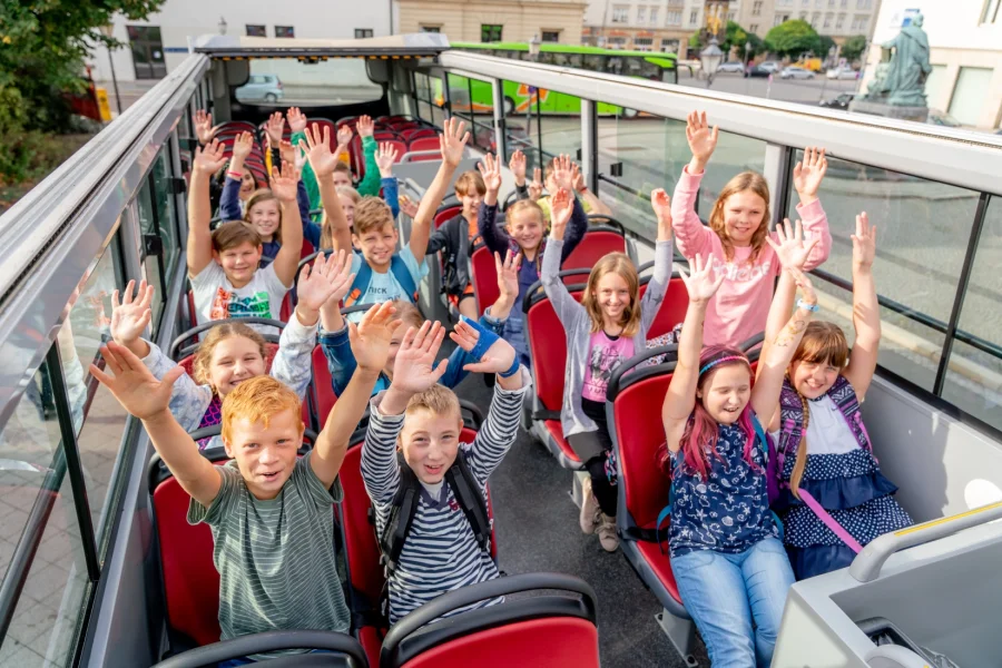 Froehliche Kindergruppe auf dem Oberdeck des Doppeldeckerbusses: alle strecken laechelnd die Arme in die Luft, im Hintergrund der Platz mit Reiterdenkmal