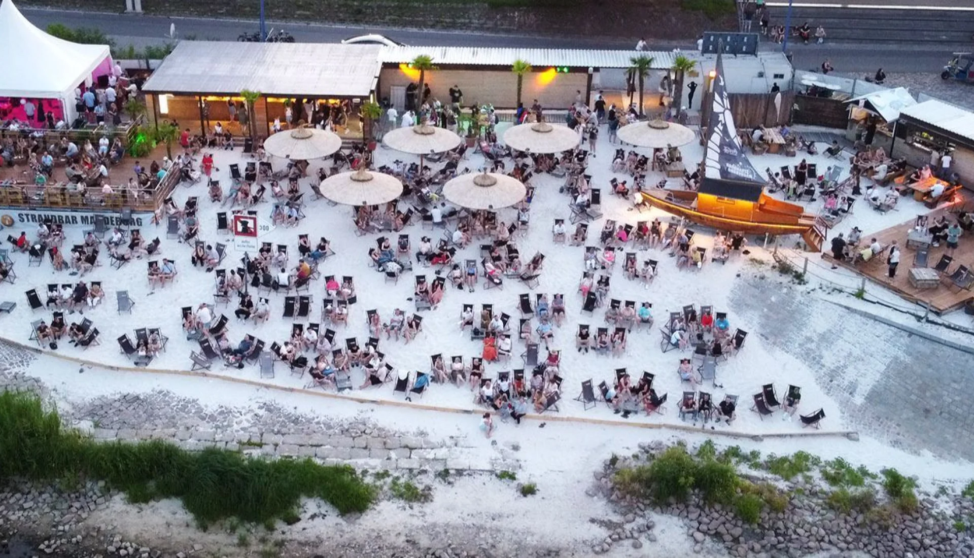 Strandbar Magdeburg aus der Luft am Abend: viele Liegest&uuml;hle im Sand, Strohschirme, beleuchtete Holzterrassen und ein Holzboot mit Segelmast