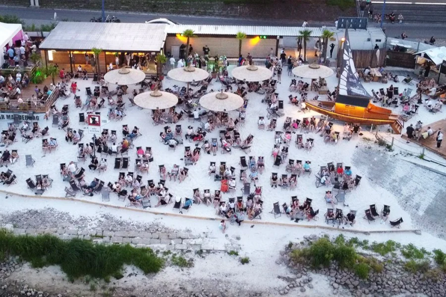Strandbar Magdeburg aus der Luft am Abend: viele Liegest&uuml;hle im Sand, Strohschirme, beleuchtete Holzterrassen und ein Holzboot mit Segelmast
