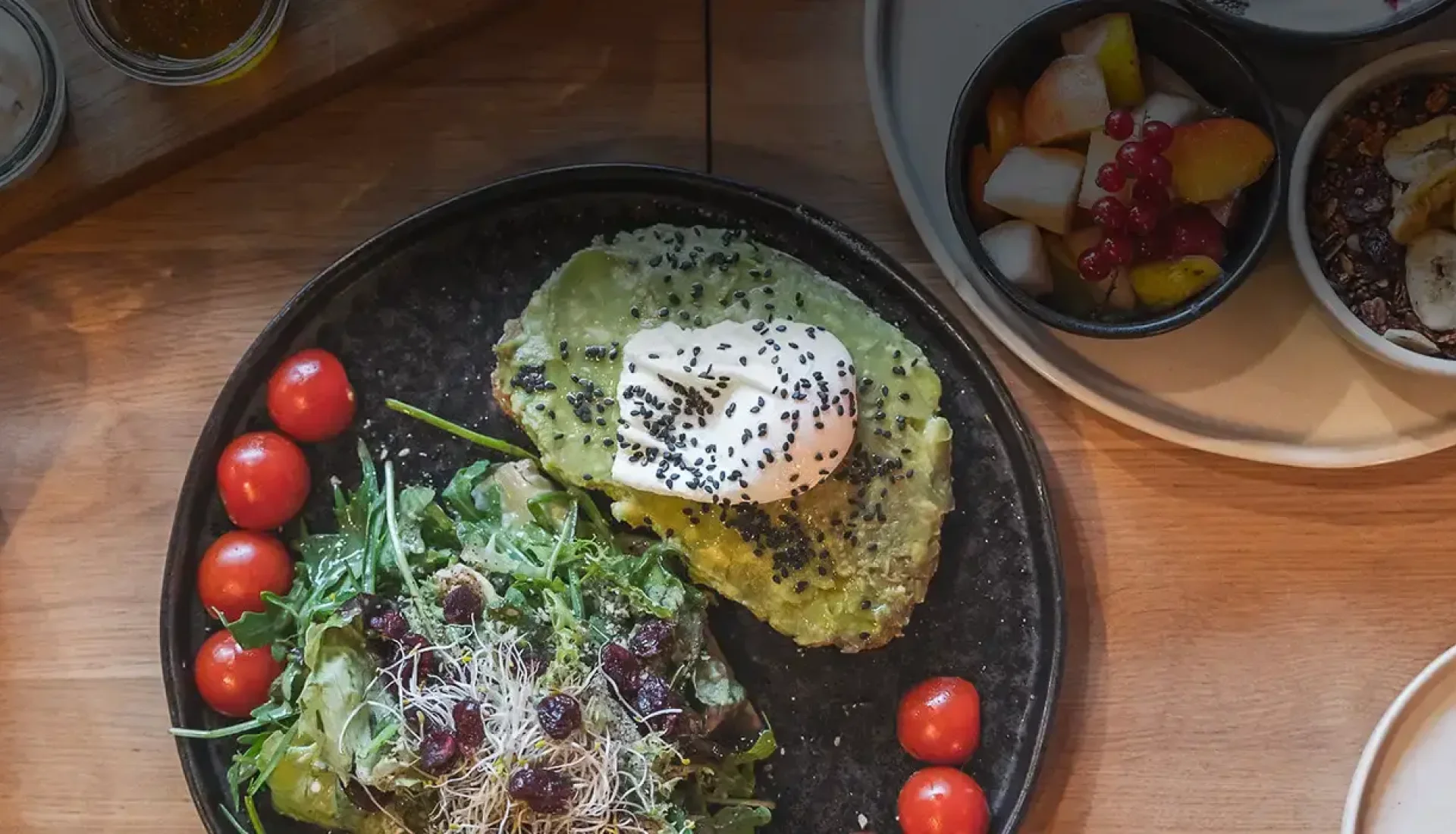 Fr&uuml;hst&uuml;ck im Thies von oben: Teller mit Avocadobrot, pochiertem Ei und Salat, dazu Obstschale, M&uuml;sli, Orangensaft und kleine Gl&auml;ser mit Aufstrichen