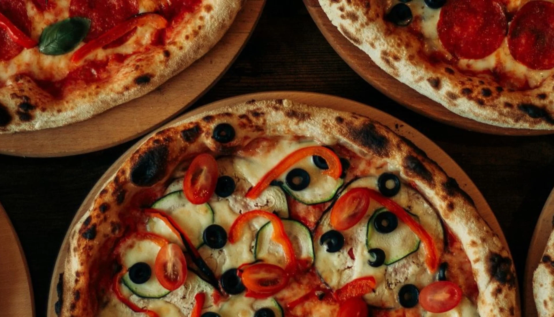 F&uuml;nf Steinofenpizzen auf runden Holzbrettern von oben, belegt mit Salami, Paprika, Oliven, Zucchini und K&auml;se