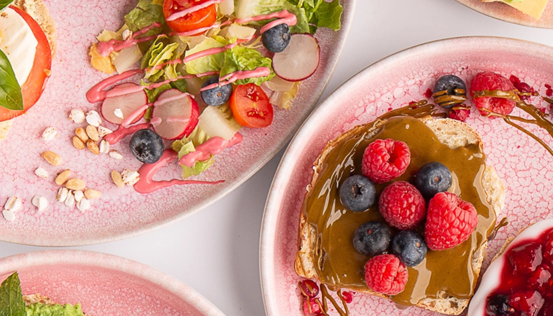 Fruehstuecksteller auf rosa Keramik im Vanillekind: Brote mit Tomate-Mozzarella-Basilikum, mit Nussmus und frischen Himbeeren und Heidelbeeren, dazu Salat mit Radieschen und Kirschtomaten