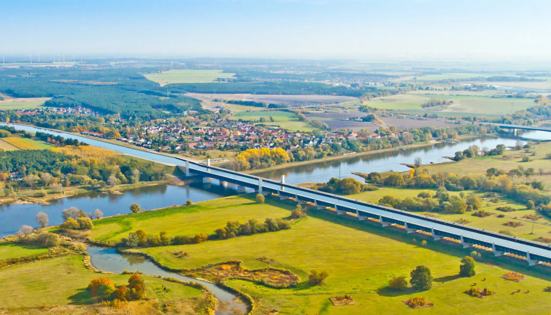 Weites Luftbild des Wasserstra&szlig;enkreuzes im Herbst: die lange Trogbr&uuml;cke quert die Elbe, dahinter Felder und ein Dorf