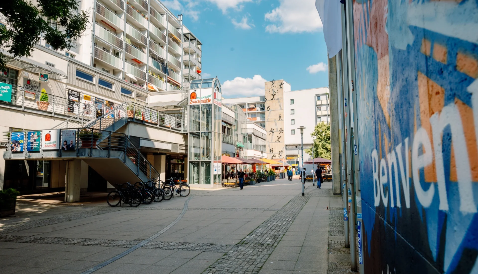 Fu&szlig;g&auml;ngerpassage am Ulrichhaus mit dem gl&auml;sernen Aufzugsturm der Magdeburger Zwickm&uuml;hle, Fahrradst&auml;ndern und Sonnenschirmen der Gastronomie