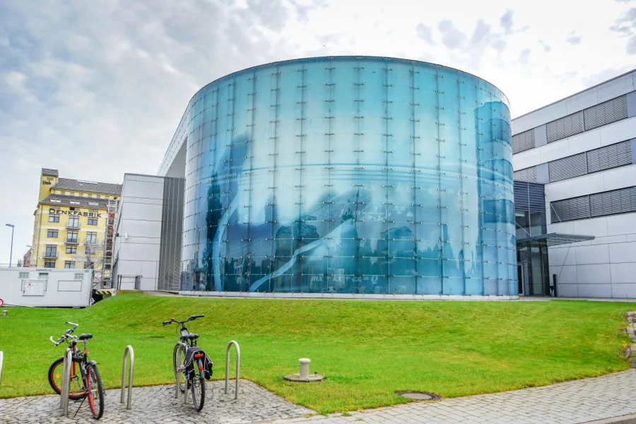 Der gl&auml;serne Rundbau des ifak am Wissenschaftshafen Magdeburg, davor eine Rasenfl&auml;che mit abgestellten Fahrr&auml;dern, links das gelbe Geb&auml;ude Denkfabrik
