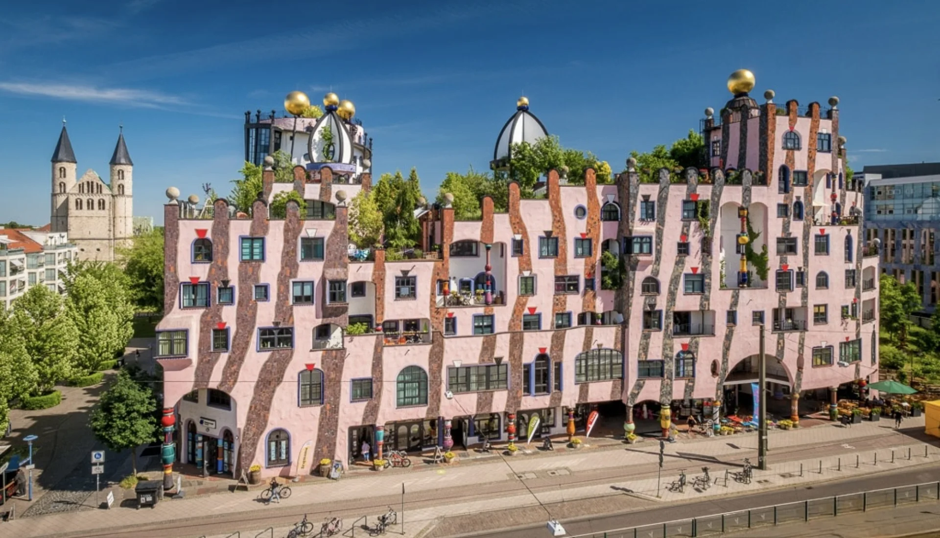 Die Gruene Zitadelle von Magdeburg: rosafarbenes Hundertwasser-Haus mit goldenen Kugeln, begruenten Daechern und bunten Keramiksaeulen, links die Tuerme des Klosters Unser Lieben Frauen