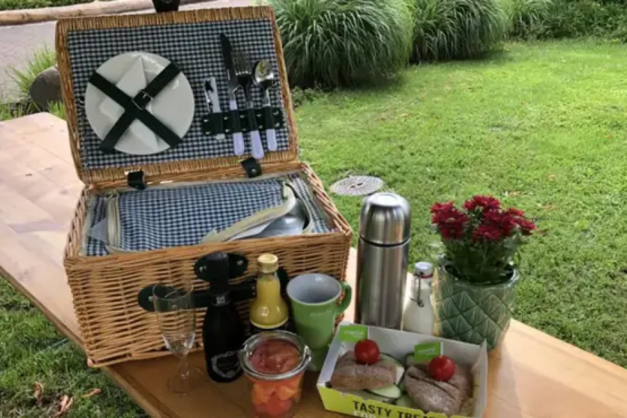 Geoeffneter Picknickkorb mit Geschirr und Besteck auf einem Holztisch im Gruenen, davor Thermoskanne, Blumen und belegte Broetchen