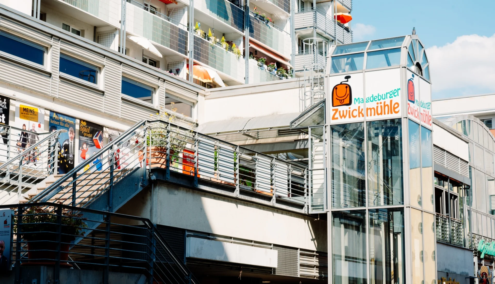 Glaeserner Aufzugsturm mit dem orangefarbenen Logo der Magdeburger Zwickmuehle vor einem Plattenbau, links die Freitreppe mit Kabarett-Plakaten