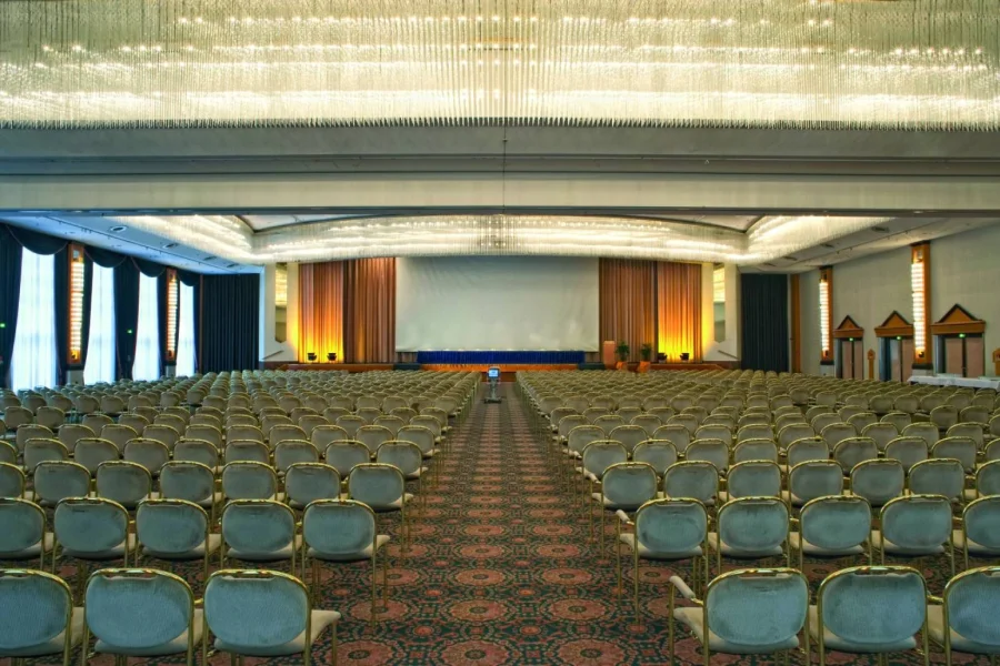 Gro&szlig;er Saal in Reihenbestuhlung mit mehreren hundert St&uuml;hlen, B&uuml;hne mit Leinwand und langen Kristallleuchtern an der Decke