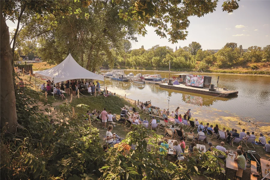 Gr&uuml;ne Wiesen mit Menschen, die auf Liegest&uuml;hlen sitzen. Im Hintergrund die Elbe mit Booten und einer Videowand auf dem Steg.