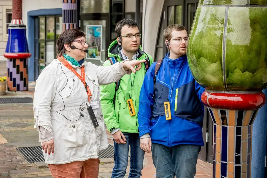 Stadtfuehrerin mit Headset zeigt zwei Gaesten mit Fuehrungsempfaengern eine gruene Keramiksaeule an der Gruenen Zitadelle
