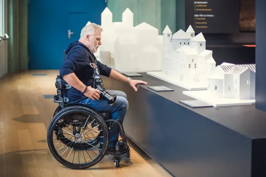 Rollstuhlfahrer mit Kamera betrachtet im Dommuseum Ottonianum weisse Architekturmodelle mittelalterlicher Kirchenbauten in einer Vitrinennische