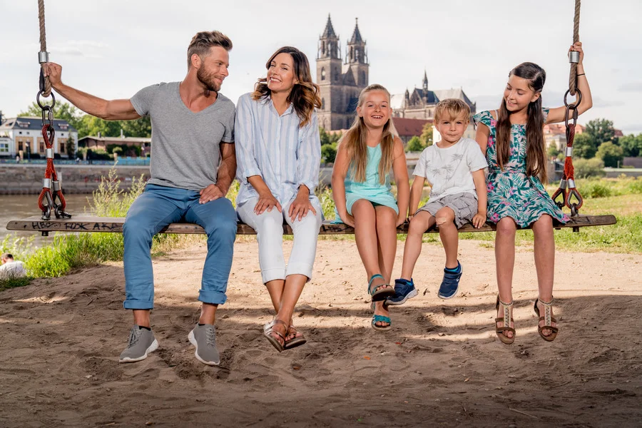 Familie mit drei Kindern sitzt lachend auf einer grossen Holzschaukel am sandigen Elbufer, dahinter der Magdeburger Dom