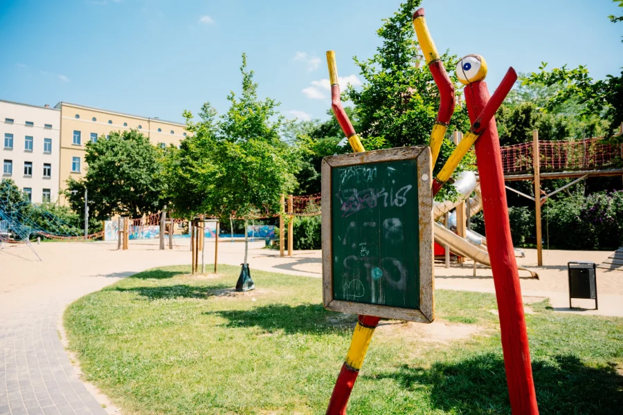 Abenteuerspielplatz Hegelstrasse mit roten Holzfiguren, gruener Tafel, Kletternetzen und Rutschen zwischen Baeumen