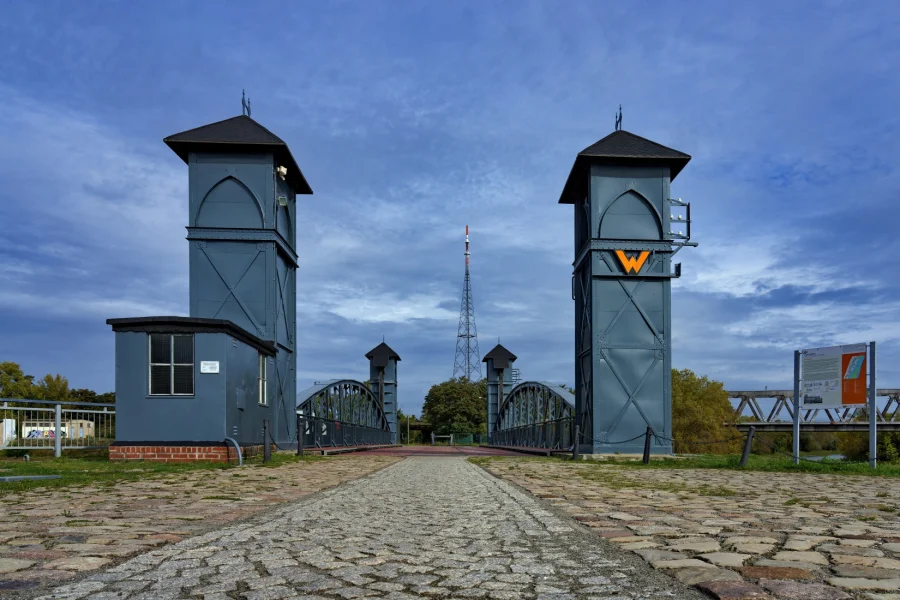 Die beiden grauen Stahltuerme der Eisenbahnhubbruecke am Wissenschaftshafen, dazwischen der gepflasterte Weg ueber die Bruecke, im Hintergrund ein Funkturm