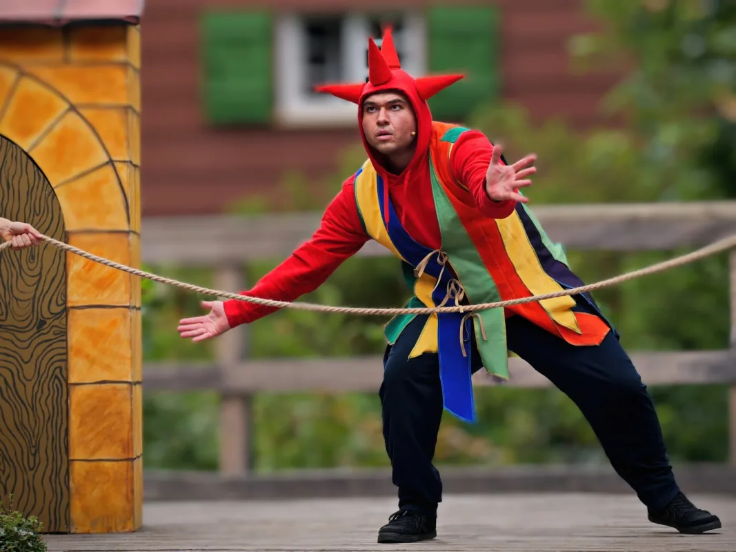 Schauspieler als Till Eulenspiegel in rot-buntem Narrenkostüm mit Zipfelmütze spielt auf einer Freilichtbühne an einem Seil