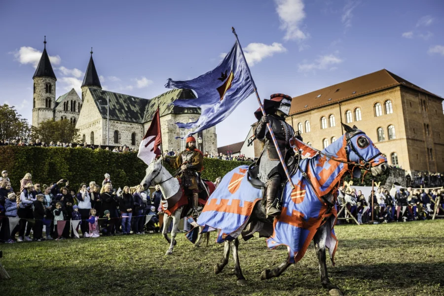 Ritter in blau-oranger Pferdedecke galoppiert mit erhobener Fahne &uuml;ber die Ritterwiese, dahinter ein zweiter Reiter, Zuschauer und die T&uuml;rme des Klosters Unser Lieben Frauen
